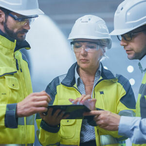 3 personnes portant des vestes techniques, des lunettes et casques de chantier en train de consulter une tablette