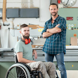 2 personnes dans un atelier, l'une en fauteuil, l'autre qui semble son tuteur se tient à ses côtés debout les bras croisés.